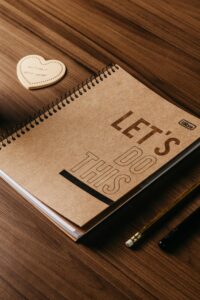 Close-up of a spiral notebook with 'Let's Do This' cover, pencil and paper heart on a wooden desk.