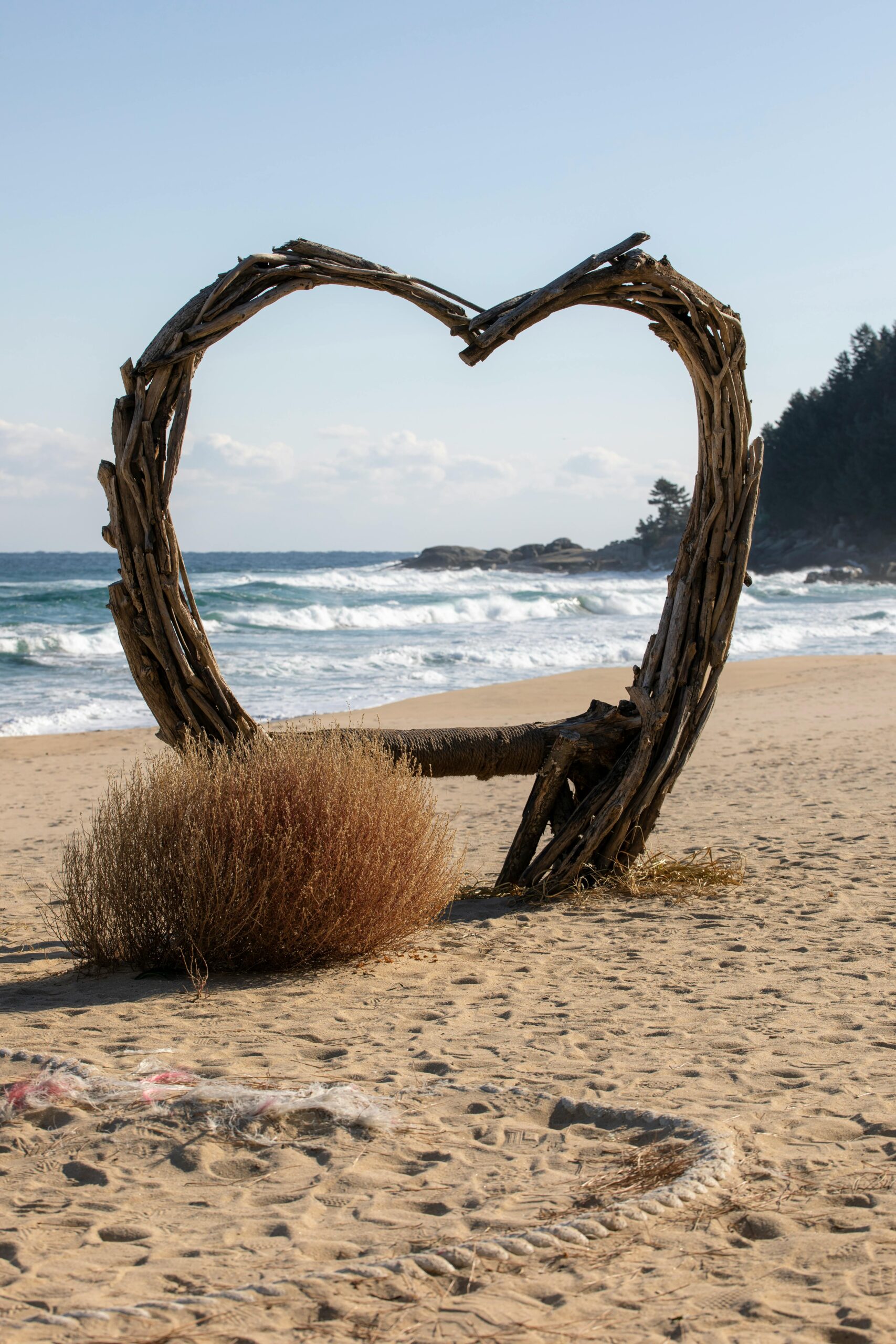 A rustic heart-shaped driftwood sculpture on a sunny Sokcho beach with waves in the background.